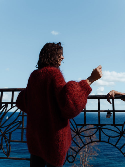 Person in a red sweater standing by a railing with a blue sky and water in the background
